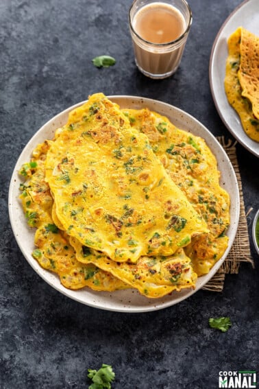 stack of besan chilla in a round white plate with the top most chilla folded and a glass of chai in the background