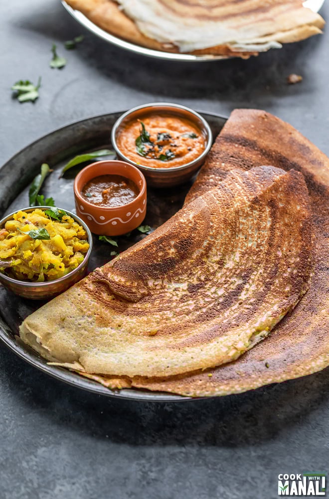 two dosa placed in a plate with bowls of chutneys and potato masala