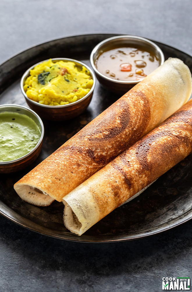 two crispy masala dosa rolled and placed in a round plate along with bowls of sambar, chutney and potato masala