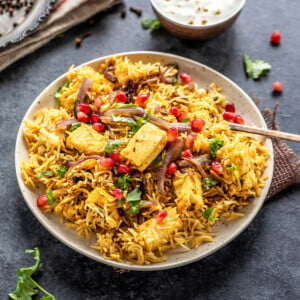 a plate full of rice and paneer, garnished with cilantro and pomegranate arils with a bowl of yogurt place on one side and a plate of spices placed on the other side