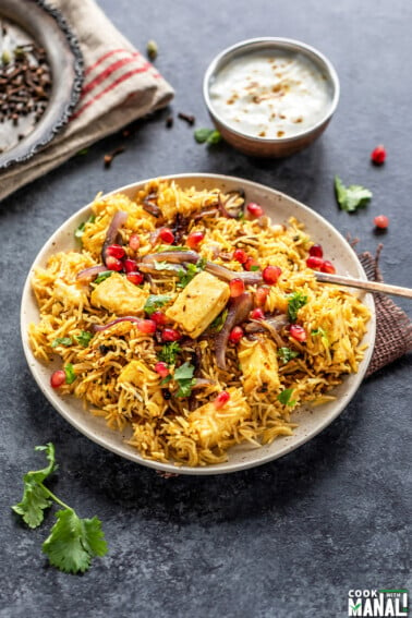 a plate full of rice and paneer, garnished with cilantro and pomegranate arils with a bowl of yogurt place on one side and a plate of spices placed on the other side