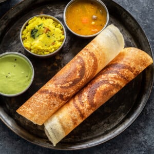 two crispy masala dosa rolled and placed in a round plate along with bowls of sambar, chutney and potato masala