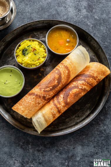 two crispy masala dosa rolled and placed in a round plate along with bowls of sambar, chutney and potato masala