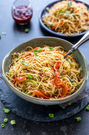 bowl of noodles with lots of veggies and another plate full of noodles in the background with a small glass jar filled with tomato ketchup