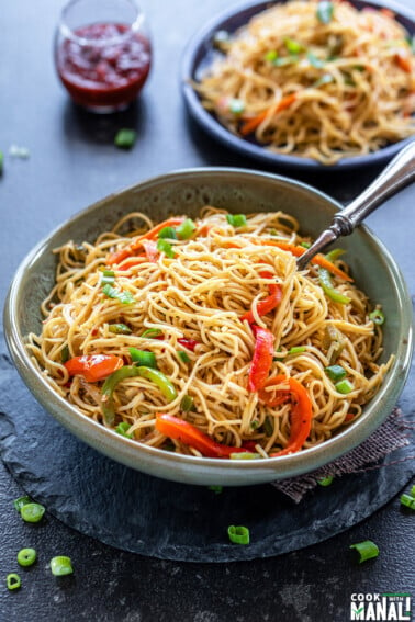 bowl of noodles with lots of veggies and another plate full of noodles in the background with a small glass jar filled with tomato ketchup