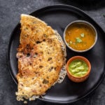 rava dosa placed in a black round plate with a bowl of sambar and a bowl of chutney