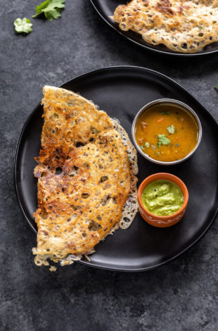 rava dosa placed in a black round plate with a bowl of sambar and a bowl of chutney