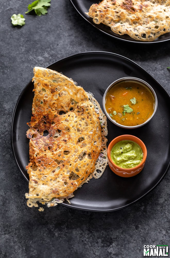 rava dosa placed in a black round plate with a bowl of sambar and a bowl of chutney