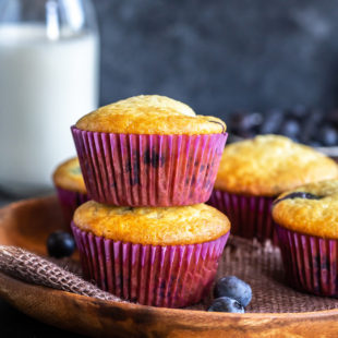 two muffins stacked on top of each other, with more muffins on the side and a bowl of blueberries and a bottle of milk in the background