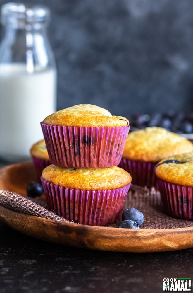 two muffins stacked on top of each other, with more muffins on the side and a bowl of blueberries and a bottle of milk in the background