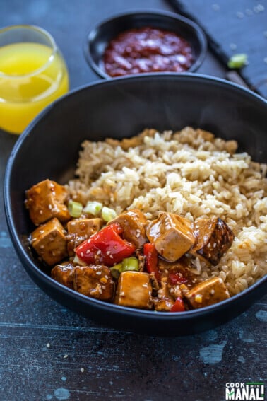 bowl of tofu topped with sesame seeds and brown rice with a glass of pineapple juice in the background with a bowl of hot sauce placed on the side