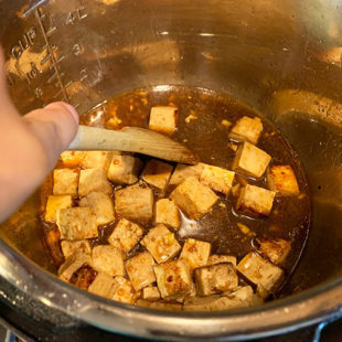 cubes of tofu with a brown color sauce and a wooden spatula scraping the bottom of the pot