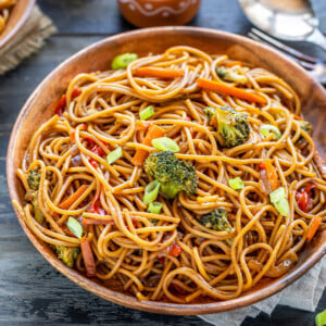 noodles with veggies served in a wooden plate with bowls of chili sauce and soy sauce placed in the background