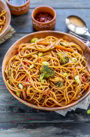 noodles with veggies served in a wooden plate with bowls of chili sauce and soy sauce placed in the background