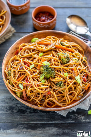 noodles with veggies served in a wooden plate with bowls of chili sauce and soy sauce placed in the background