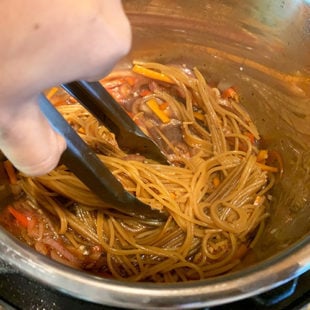 a pair of tongs holding spaghetti noodles in an instant pot