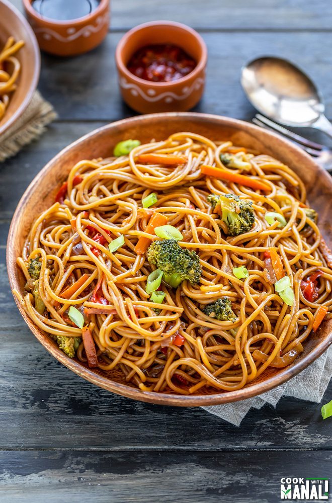 noodles with veggies served in a wooden plate with bowls of chili sauce and soy sauce placed in the background