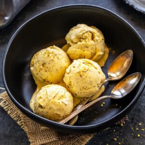 4 scoops of kesar elaichi ice cream in a black bowl served with two spoons with a plate of cardamom pods in the back and a small container of saffron on the side