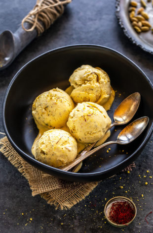 4 scoops of kesar elaichi ice cream in a black bowl served with two spoons with a plate of cardamom pods in the back and a small container of saffron on the side
