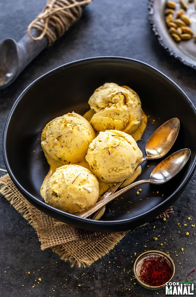 four scoops of kesar elaichi ice cream in a black bowl with two spoons and a small container of saffron placed on one side and cardamom pods in the background