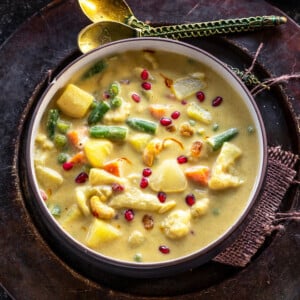 yellow color vegetable curry in a round bowl garnished with pomegranate arils and two golden color spoons placed in the background