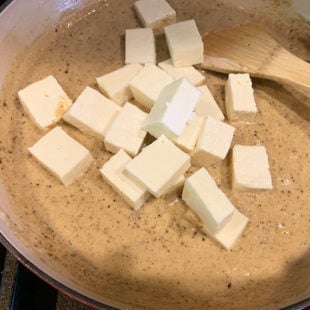 paneer cubes being added to a cream colored sauce in a pan