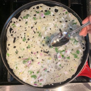 oil being drizzled all around a dosa (Crepe) on a iron pan with a spoon