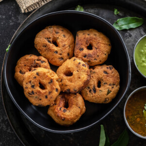 medu vada arranged in a black bowl with curry leaves placed on the side and spoons placed in the background