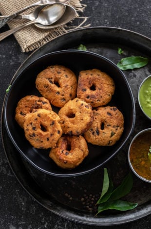 medu vada arranged in a black bowl with curry leaves placed on the side and spoons placed in the background