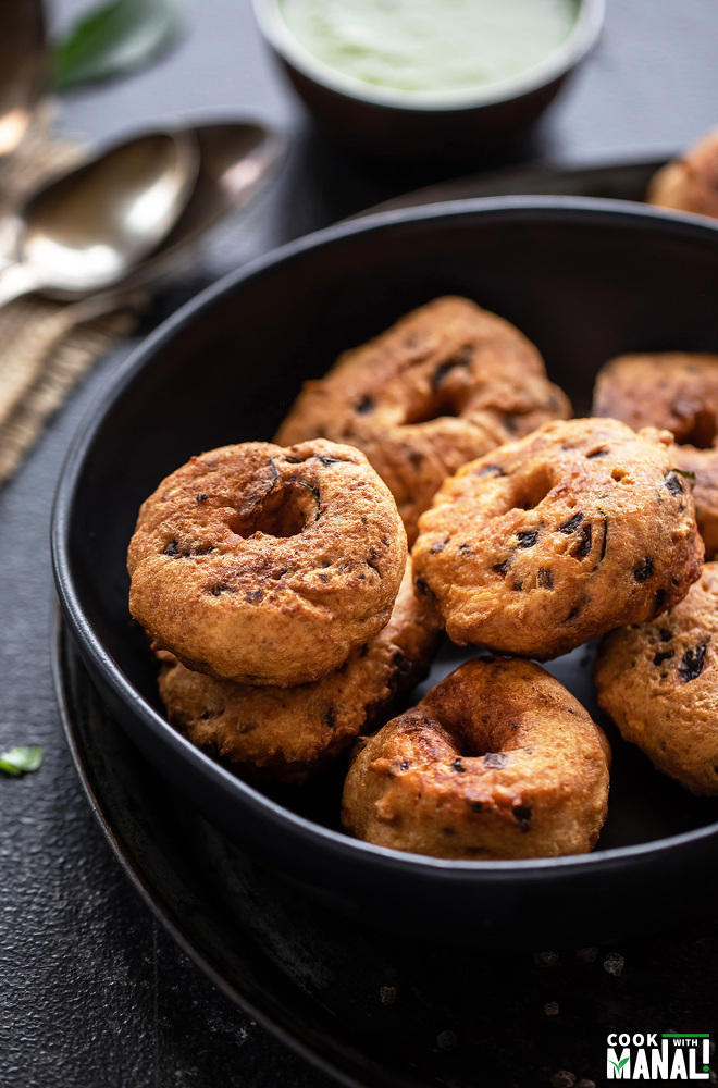 medu vada in a black bowl with bowl of chutney on the background and spoons placed on the side