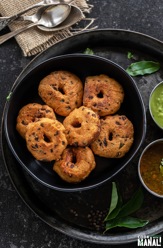 medu vada arranged in a black bowl with curry leaves placed on the side and spoons placed in the background