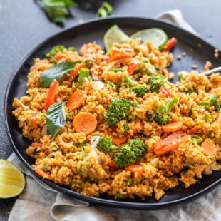 black plate with quinoa, veggies, basil, lime wedge and more basil and a bowl of hot sauce in the background