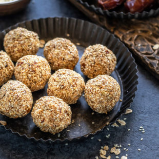 sesame oats ladoo (balls) placed in a rimmed black plate with some rolled oats scattered around and a bowl with sesame seeds in the background