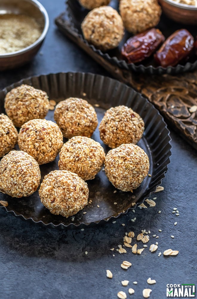 sesame oats ladoo (balls) placed in a rimmed black plate with some rolled oats scattered around and a bowl with sesame seeds in the background