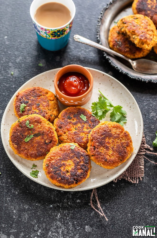 sweet pottao tikki arranged in a white plate with a small bowl of ketchup on the side a glass of chai placed in the background