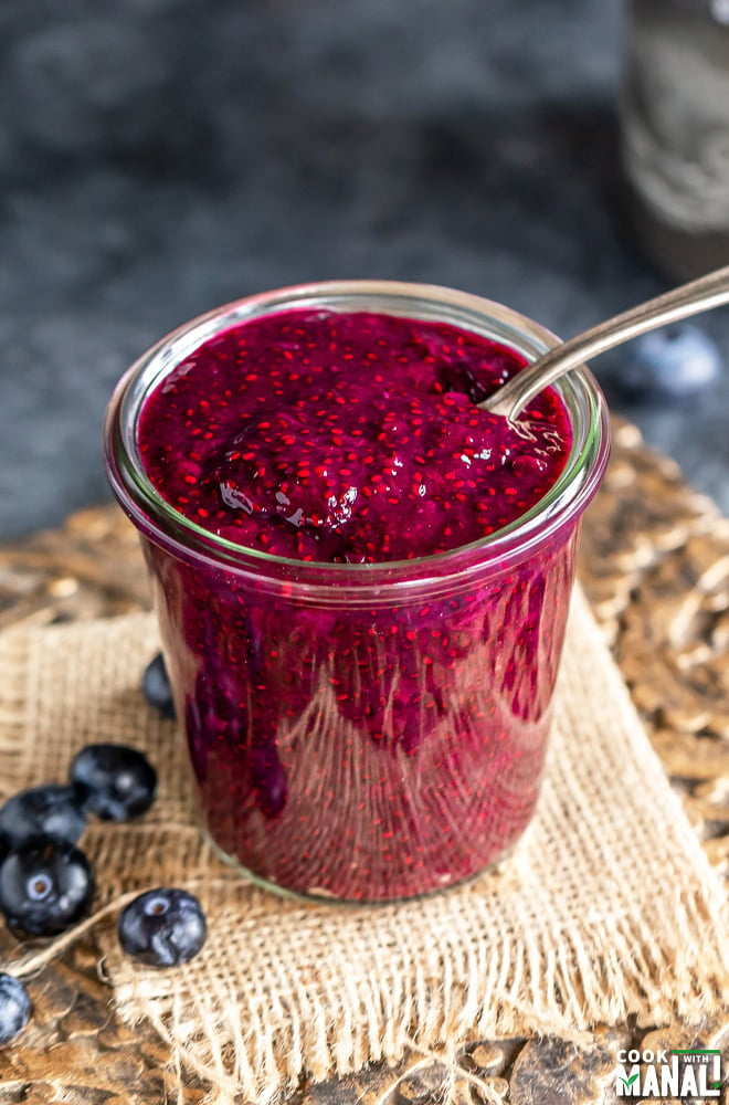 a spoon scooping out jam from a glass jar with few blueberries lying on the side of the glass jar