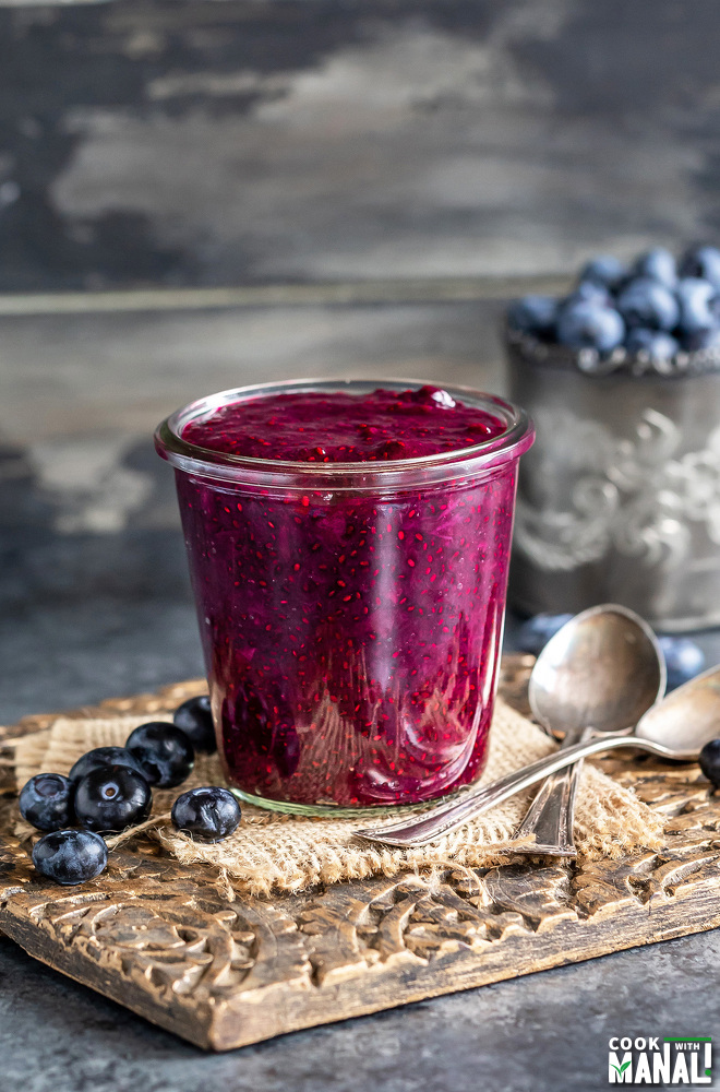 glass jar filled with blueberry chia jam with 2 spoon placed on the side and a jar of blueberries placed in the background