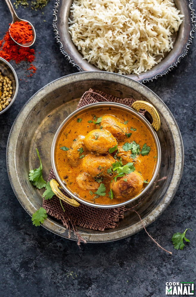 baby potato curry served in a copper kadai with a plate of rice in the background and a spoon of red chili powder placed on the side