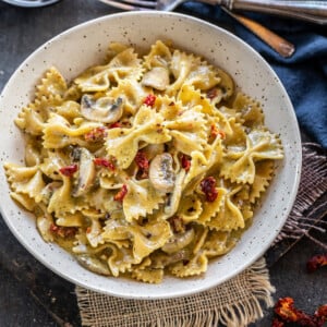 bowl of bow tie pasta with mushrooms and sun-dried tomatoes and a bowl of pesto placed in the background