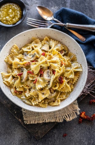 bowl of bow tie pasta with mushrooms and sun-dried tomatoes and a bowl of pesto placed in the background