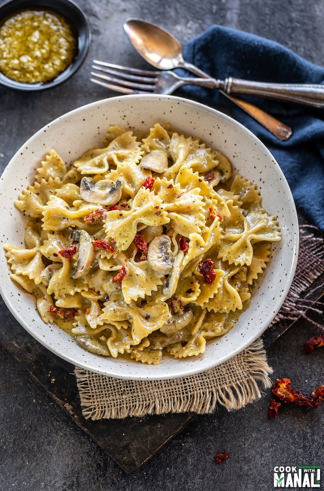 bowl of bow tie pasta with mushrooms and sun-dried tomatoes and a bowl of pesto placed in the background