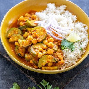 zucchini chickpea curry served with white rice in a yellow color bowl topped with sliced onion, cilantro and a lime wedge