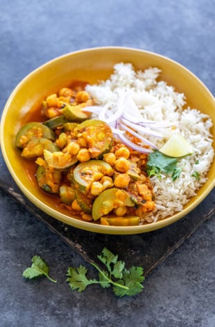 zucchini chickpea curry served with white rice in a yellow color bowl topped with sliced onion, cilantro and a lime wedge