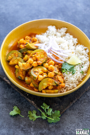 zucchini chickpea curry served with white rice in a yellow color bowl topped with sliced onion, cilantro and a lime wedge