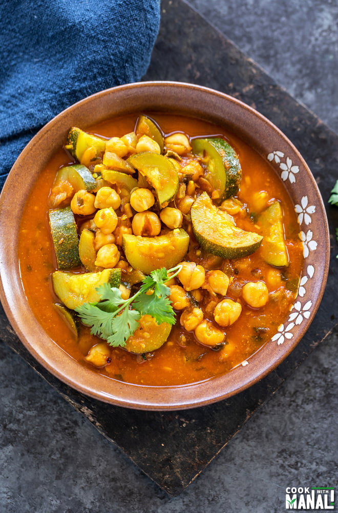 zucchini and chickpea curry served in a brow color bowl and a blue napkin placed in the background