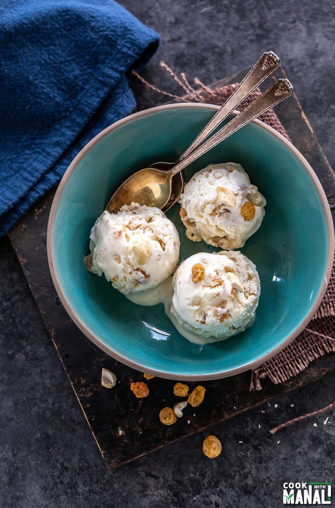 overhead shot of 3 ice cream scoops served in a blue bowl with few cashews and golden raisins scattered on the sides