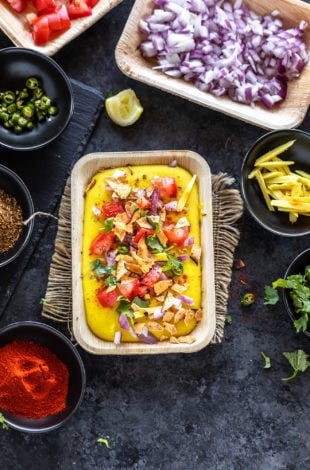 moong dal chaat served in a rectangular serving bowl topped with tomatoes, onion and bowls of cilantro, chili powder, onion, tomatoes placed in the background and on the sides
