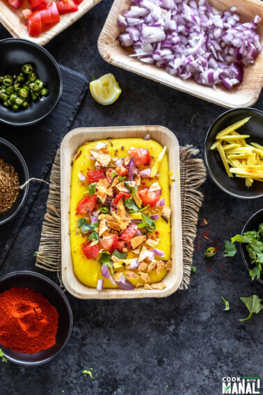 moong dal chaat served in a rectangular serving bowl topped with tomatoes, onion and bowls of cilantro, chili powder, onion, tomatoes placed in the background and on the sides