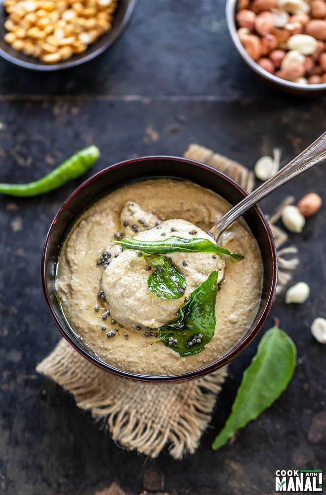 a spoon scooping out peanut chutney from a wooden bowl with few curry leaves and peanuts scattered on the sides