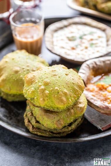 stack of puris placed in a plate along with glass of chai and bowls of curry and raita